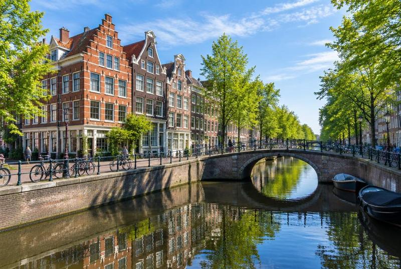 A sunlit canal in Amsterdam's Jordaan, brick canal houses and a small arched bridge reflected in still water.