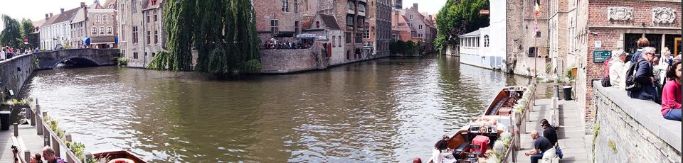 Weiter Blick auf die Biegung der Brügger Gracht, mit Backsteinhäusern, einer Weide und Ausflugsbooten am Kai.
