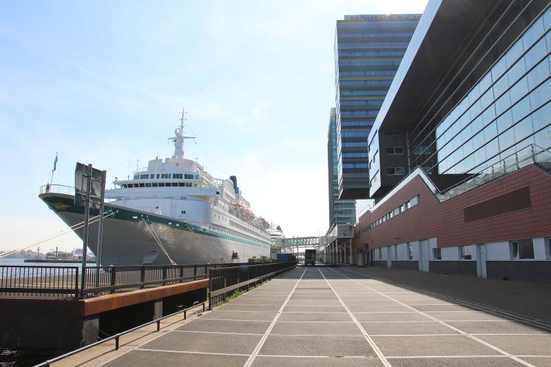 A white cruise ship moored alongside the quay of the Passenger Terminal Amsterdam on a sunny day.