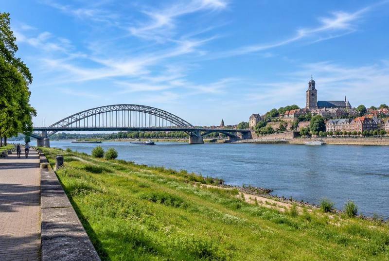 The Waal at Nijmegen on a clear day, a steel arch bridge and the town rising on the hill beyond the river.