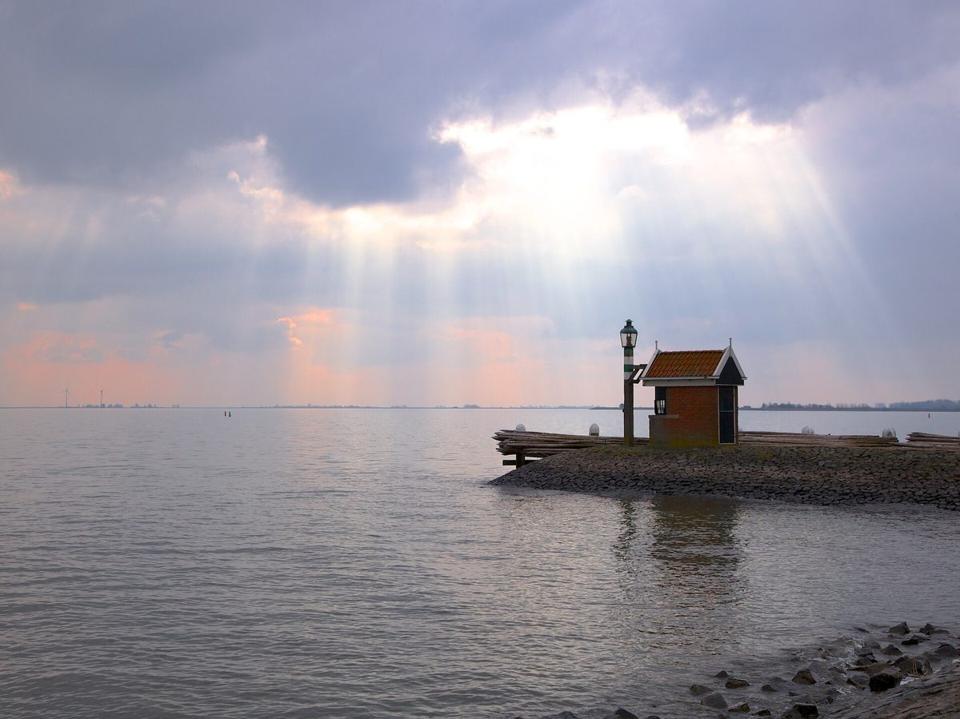 Ein kleines gemauertes Hafenfeuer auf einer Mole, während Sonnenstrahlen über dem IJsselmeer durch die Wolken brechen.
