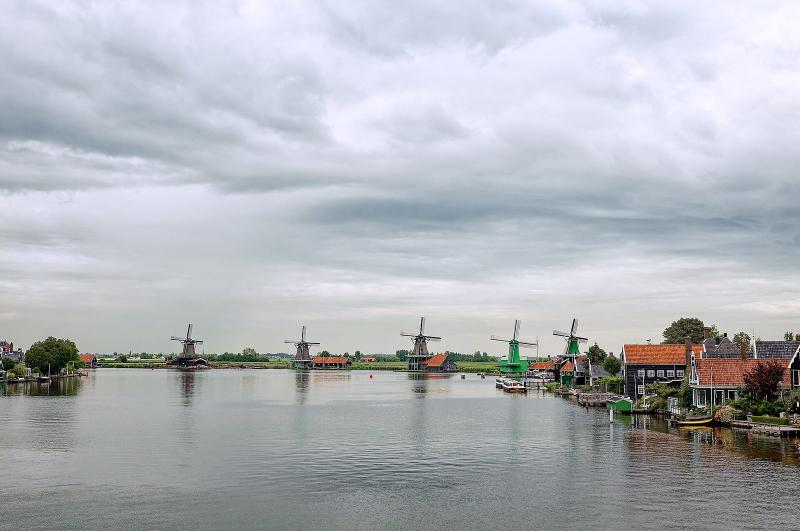 Five working windmills standing in a row along the river Zaan, with green wooden houses on the bank beside them.