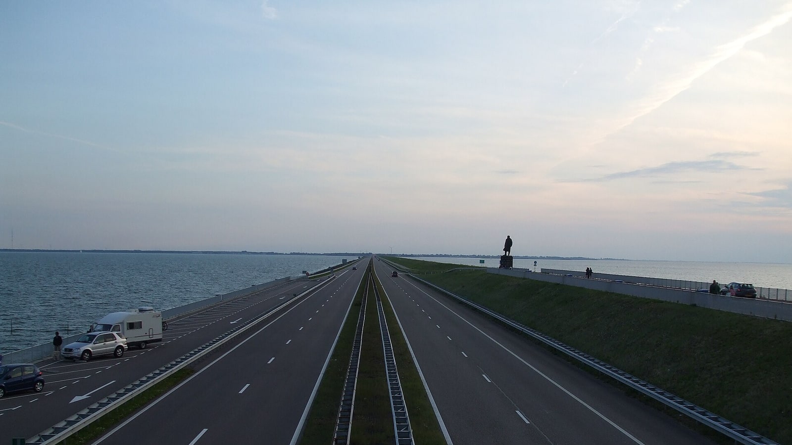 The motorway running dead straight along the Afsluitdijk at dusk, water on both sides and a statue silhouetted on a plinth beside the road.