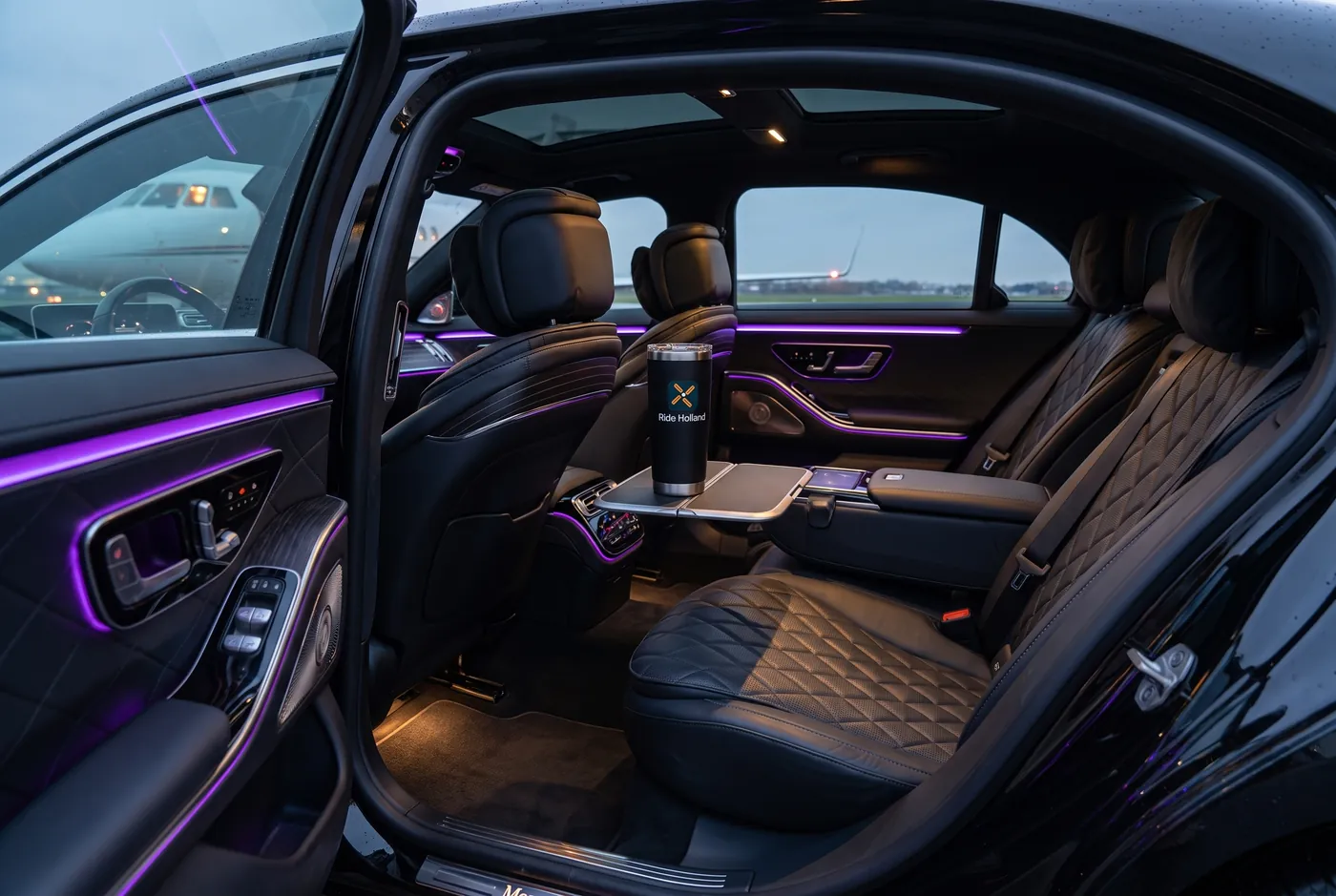 L’abitacolo posteriore di una Mercedes Classe S in pelle trapuntata nera, luce viola lungo le modanature e una tazza Ride Holland sul tavolino, con un jet oltre il finestrino.