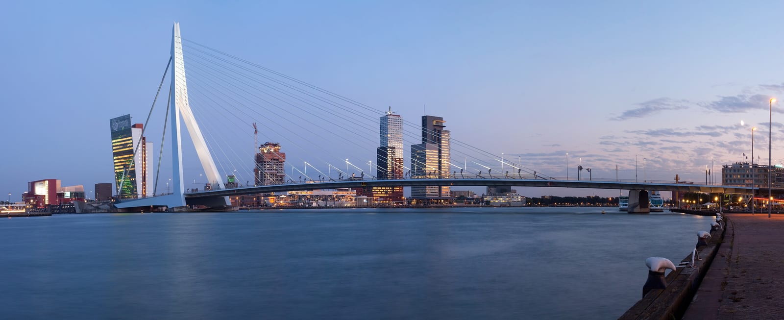 The Erasmus Bridge across the Maas at dusk, with the towers of the Kop van Zuid lit behind it and the river wide and still in the foreground.