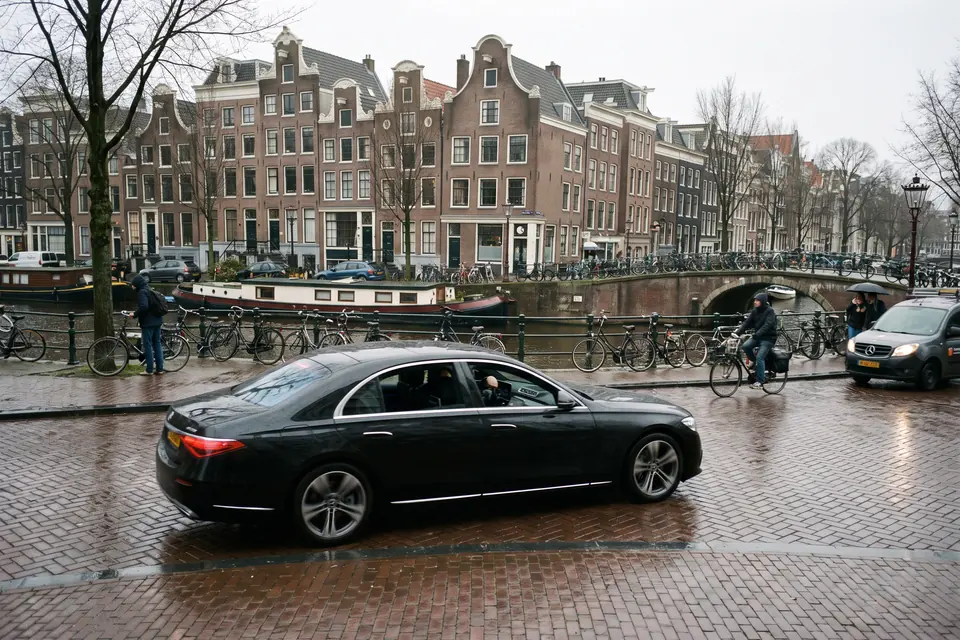 Una berlina Mercedes nera percorre una strada bagnata lungo un canale di Amsterdam, tra case con i frontoni, case galleggianti e biciclette legate alla ringhiera.