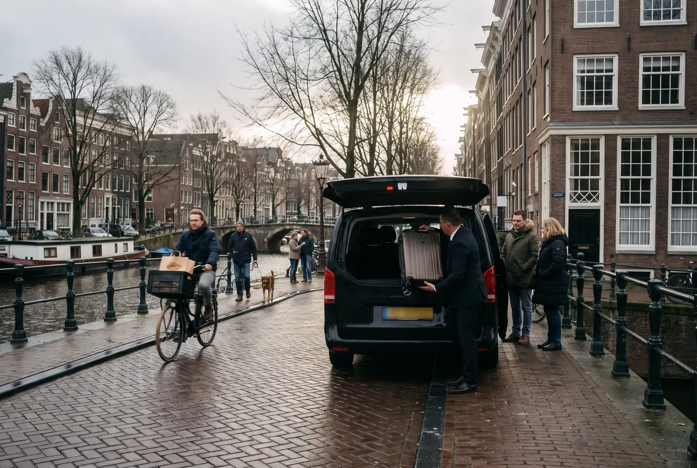 Lungo un canale di Amsterdam, un autista solleva una valigia dal portellone aperto di una Mercedes Classe V nera mentre una coppia attende sul selciato, accanto alle biciclette.