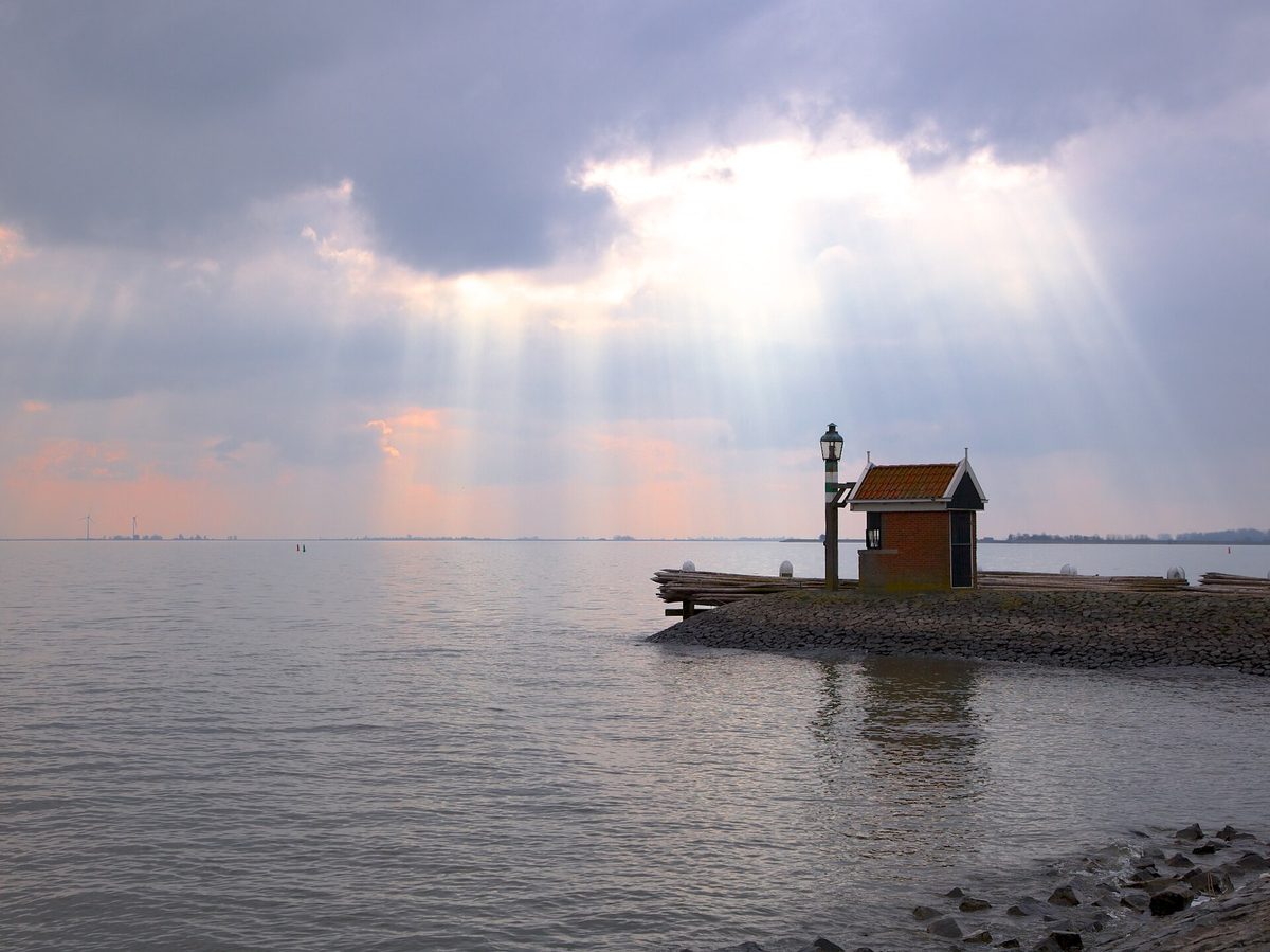 Een klein bakstenen havenlicht op een stenen strekdam, met zonnestralen door de wolken boven het IJsselmeer.